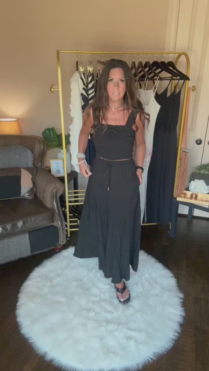 A woman viewed from behind wearing a black ruched spaghetti-strap dress, showing the back of the garment in an indoor room setting with a clothing rack visible in the background.
