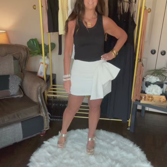 Woman in a black top and white skirt with a veil standing on a white rug in a room with a clothing rack and furniture.