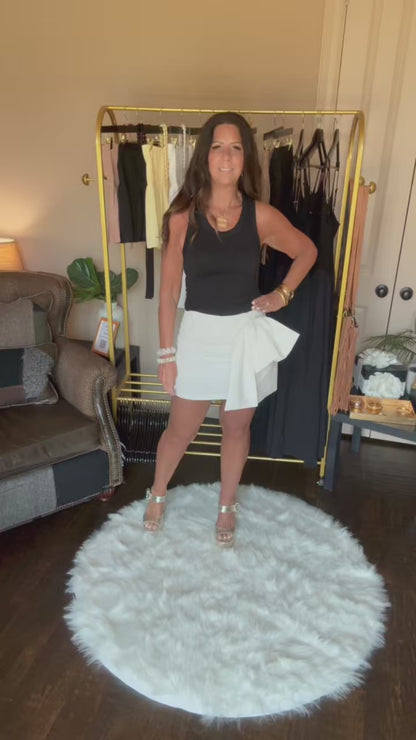 Woman in a black top and white skirt with a veil standing on a white rug in a room with a clothing rack and furniture.