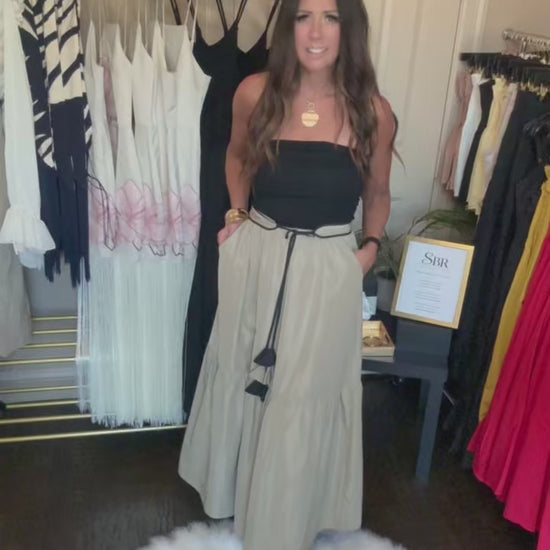 A woman modeling a long, light beige trench coat over a black top in a boutique setting, with clothing racks and a white faux fur rug visible.