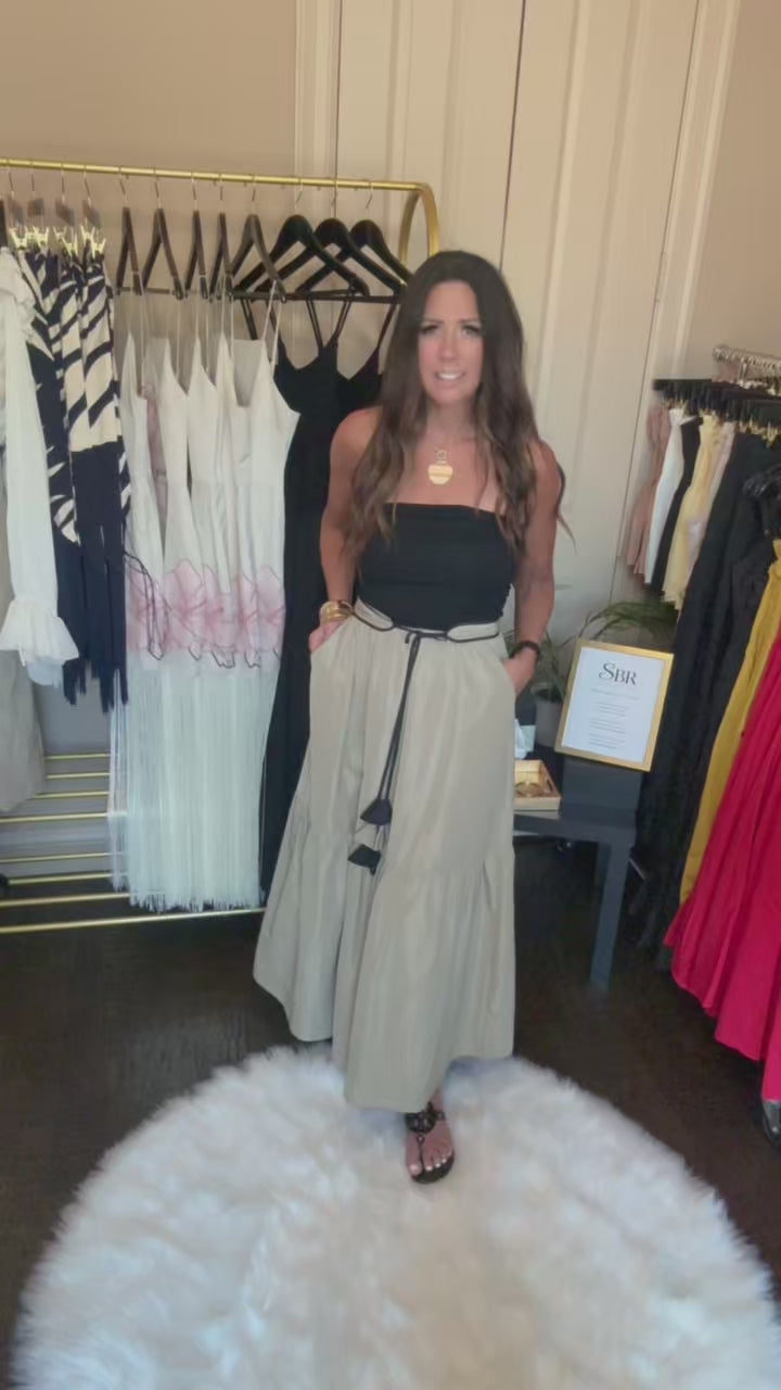 A woman modeling a long, light beige trench coat over a black top in a boutique setting, with clothing racks and a white faux fur rug visible.