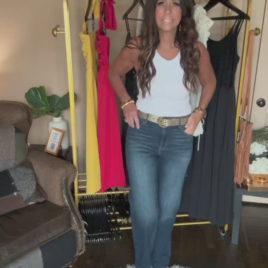 Woman standing in a room with a clothing rack and various garments modeling a pair of dark denim jeans and a white tank.